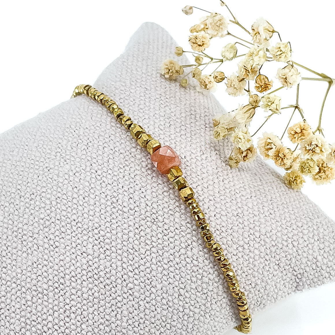 Bracelet mettant en lumière une perle cubique facettée de pierre fine naturelle de Jaspe rose pêche. Elle est entourée de perles d'hématites dorées. 